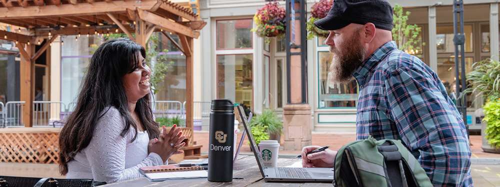 Two CU Denver students sitting opposite of each other at a picnic table with a laptop and CU Denver water bottle between them