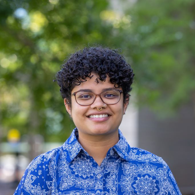 Maitreyi is a South Asian person with short, dark curly hair and glasses. She is wearing a blue button-down shirt with a white paisley pattern and is smiling.
