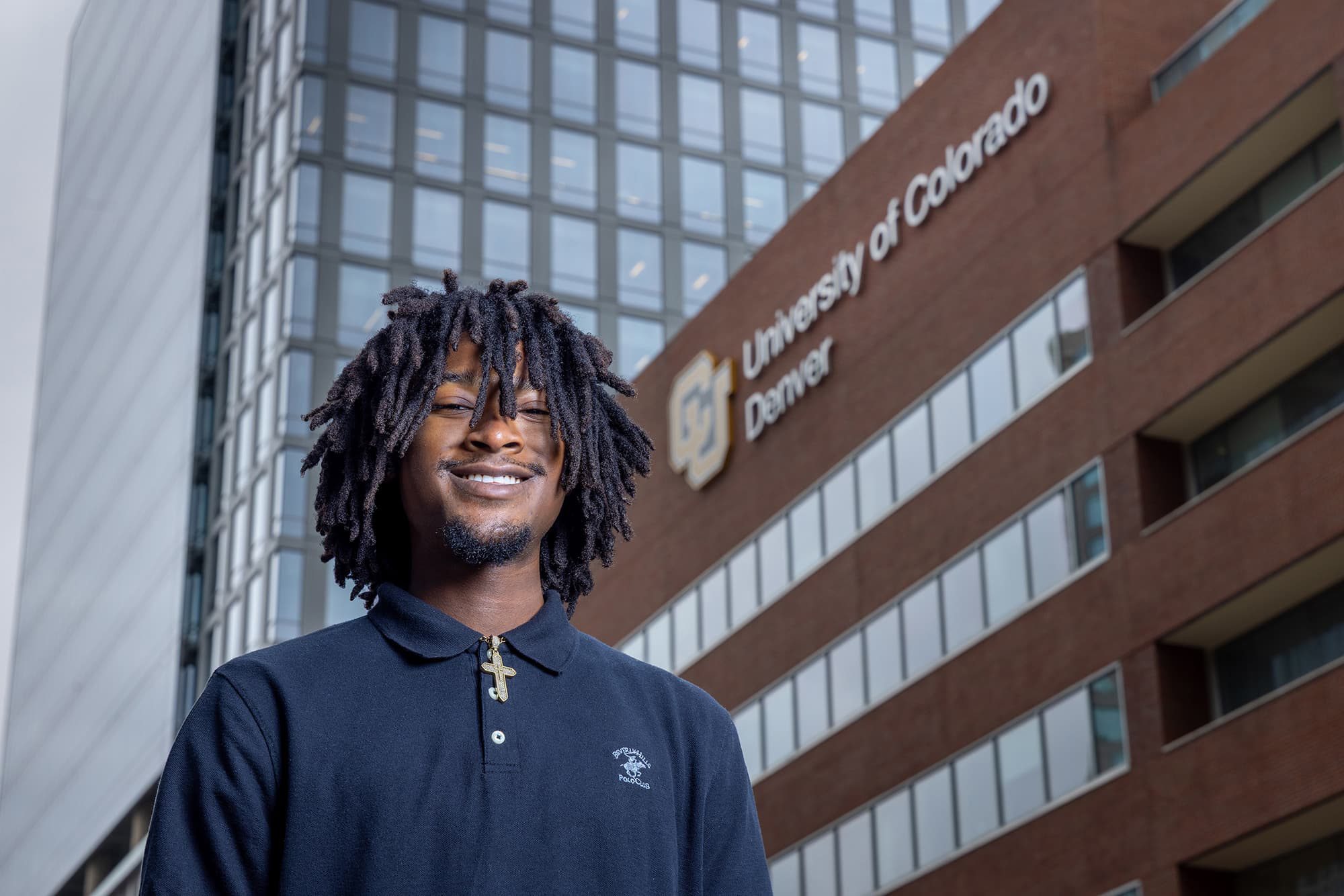 Man in a dark polo smiling with his teeth, the CU Denver Business School is in the background.