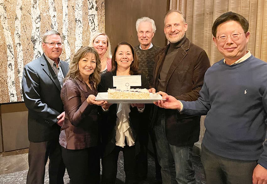 Group of people smiling around a cake with an oversized check sticking up from the cake.