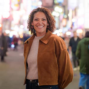 A woman wearing a tan jacket with shoulder-length dark curly hair smiling with her teeth.