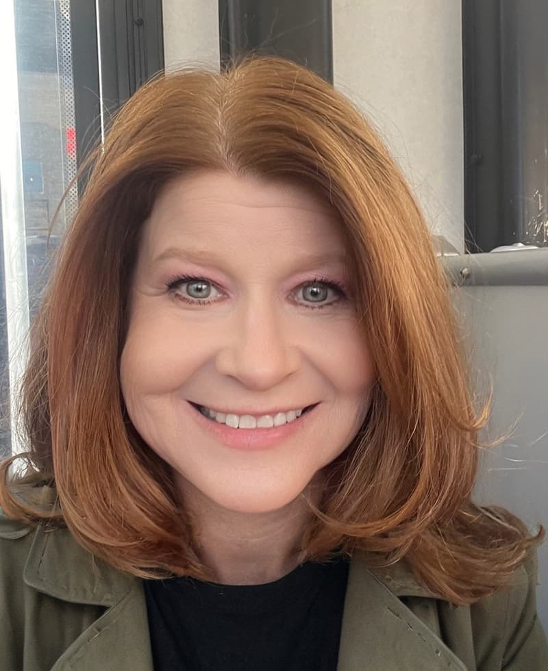 Close up of a woman's face with shoulder-length red hair and smiling with her teeth.