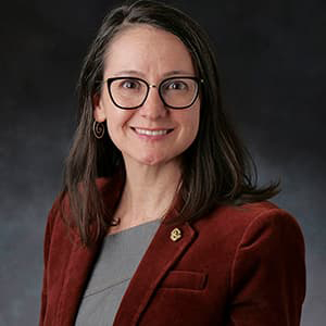 Photo of Susan Cleveland with shoulder-length dark hair, dark-rimmed glasses, wearing a deep red suit jacket, and smiling with her teeth.