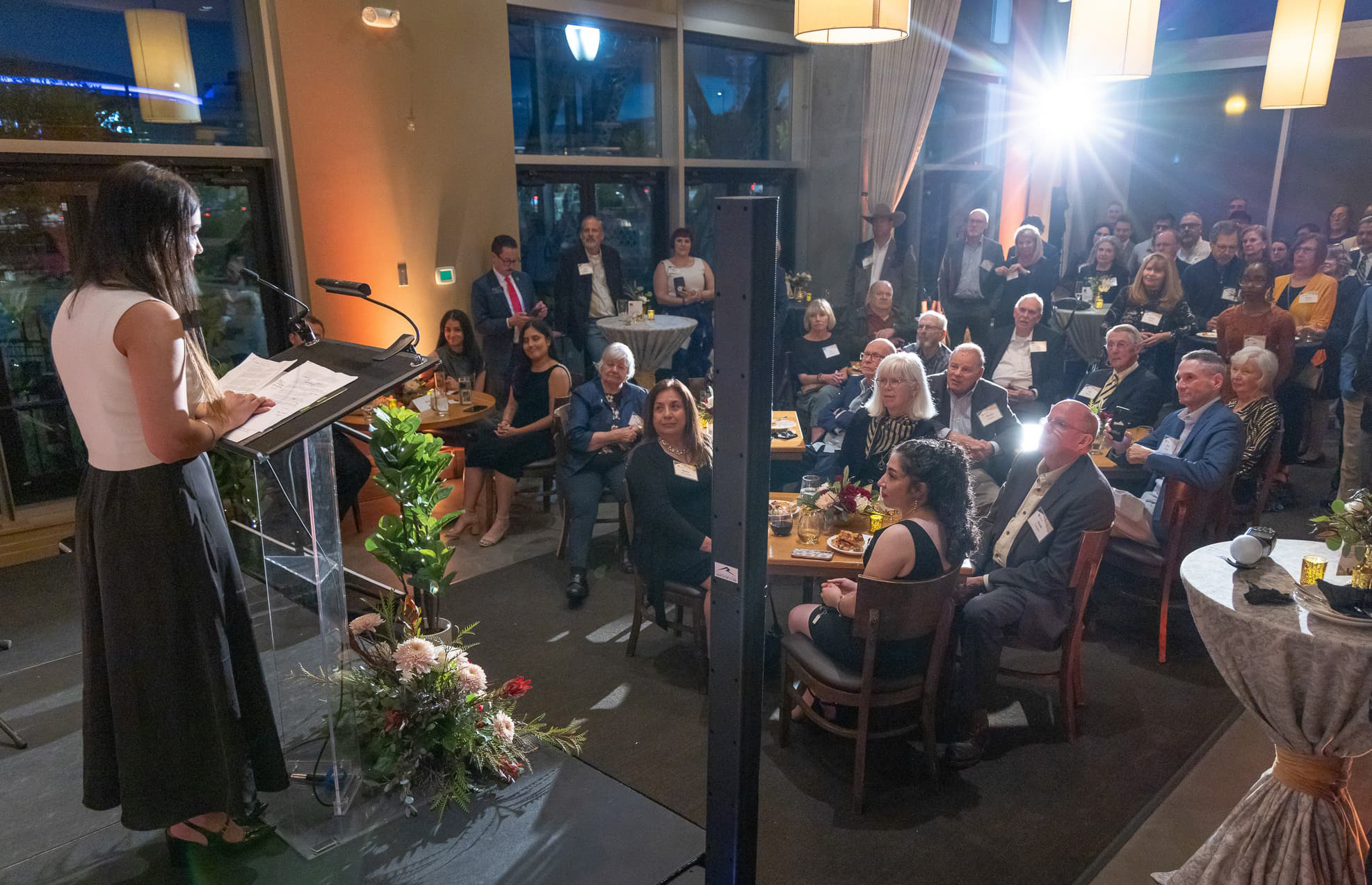 View from stage of a speaker at a podium speaking to a large crowd dressed in business attire.