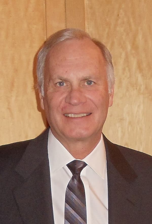 Andrew Kenney wearing a suit and tie with sort white hair and smiling with his teeth. 
