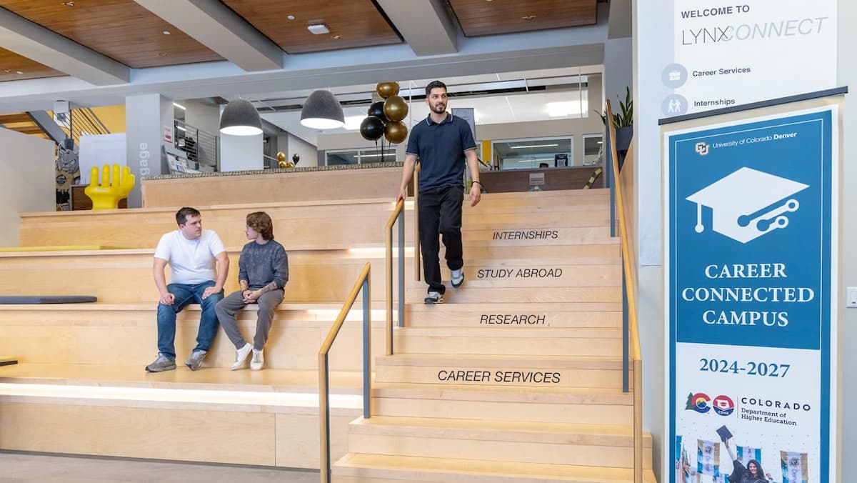 Light colored oversized wood stairs at the entrance of LynxConnect with two students seated and engaging while another walks down the stairs.