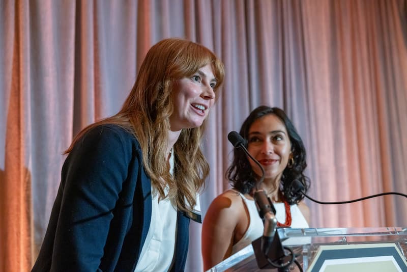 Two women dressed in professional attire speak at a podium and smile.
