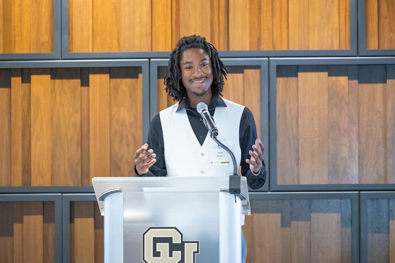 Man in a black collard shirt and white vest with shoulder-length black locks speaks at a lectern smiling and using hand gestures. 