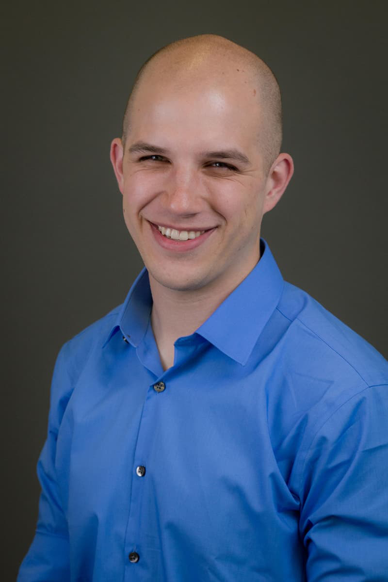 A bald Jacob Altholz in a blue button up shirt, smiling with his teeth.