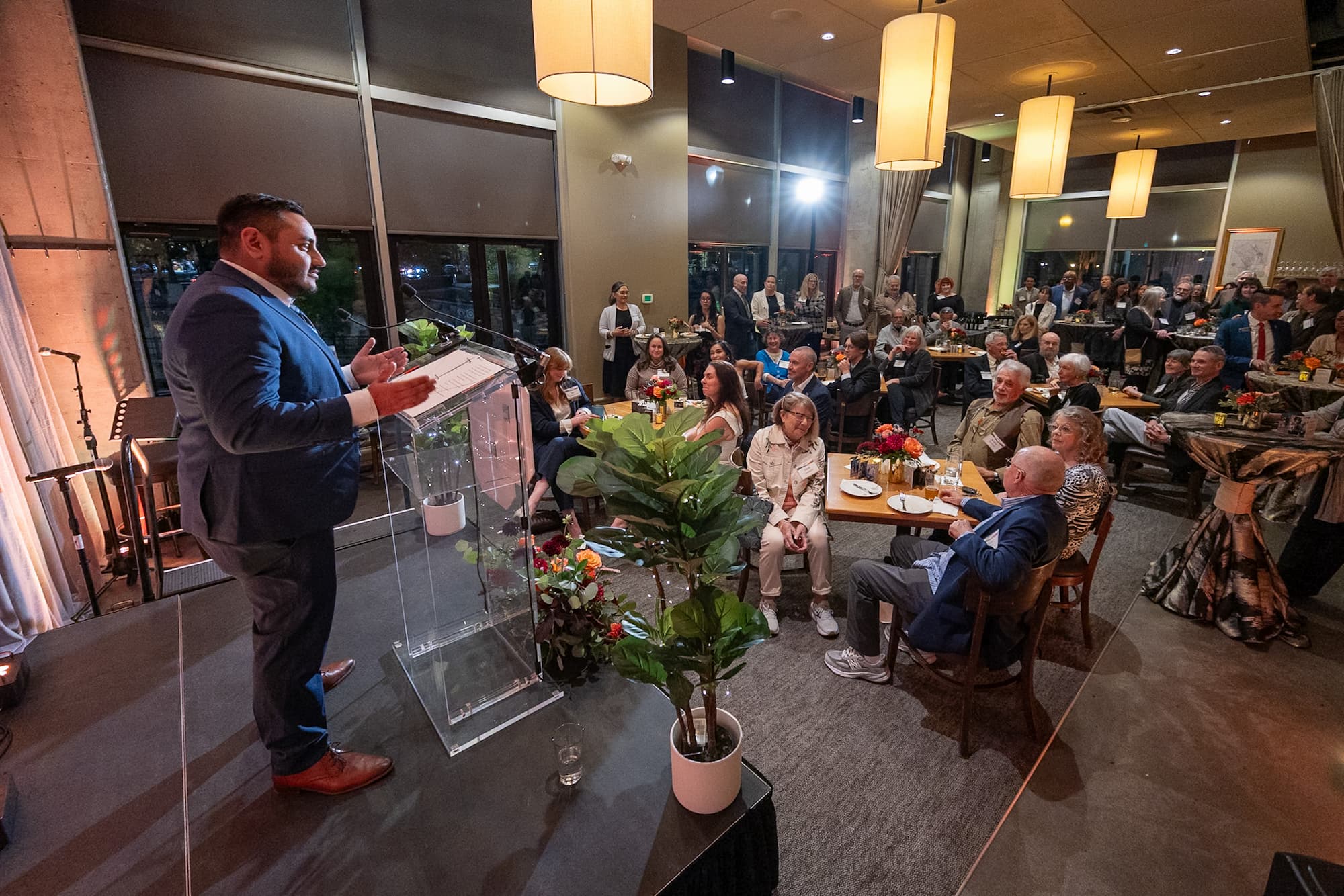 Man at a podium in a suite speaks to a cozy crowd.