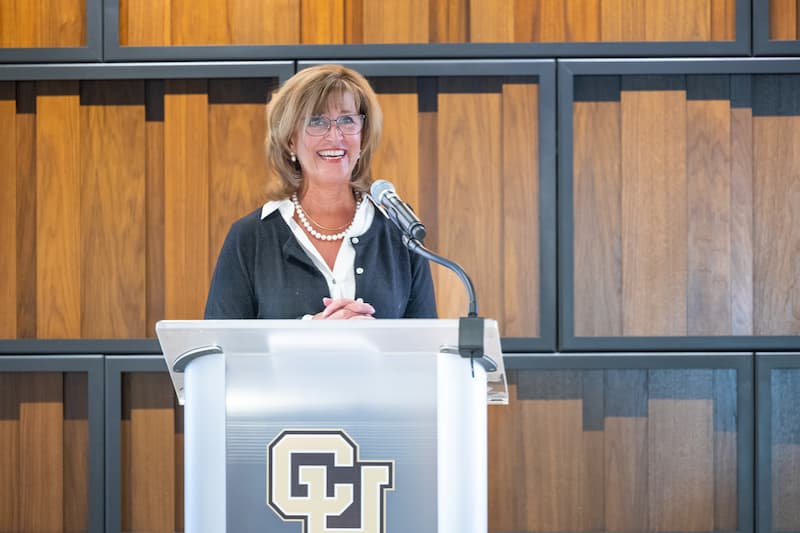 Woman wearing business attire with shoulder-length blonde hair and glasses, smiles big at a lecturn. 