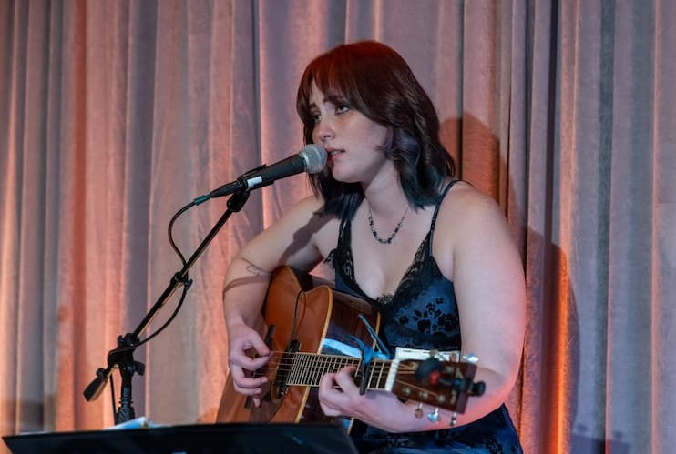 Woman with dark shoulder-length hair sits and plays a guitar while singing into a microphone.