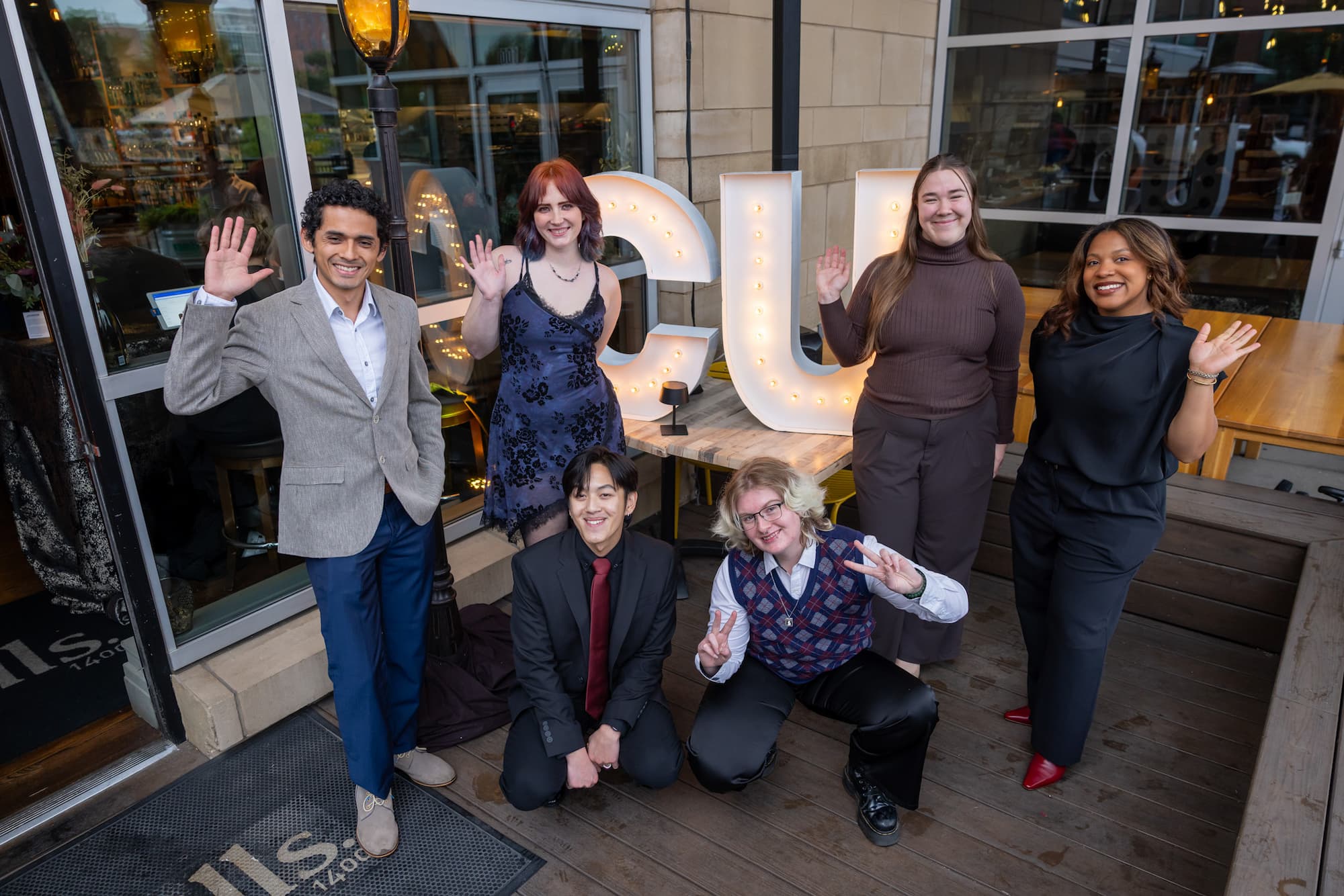Six students dressed in business attire pose together smiling and waving. 