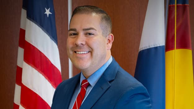Man in a blue suit and red tie smiles with his teeth in front of an American flag.