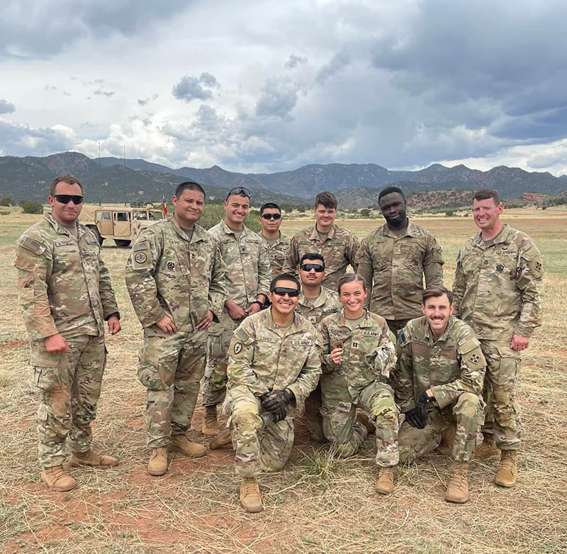 Group of 11 U.S. Army personnel smiling outdoors with mountains in the background. 