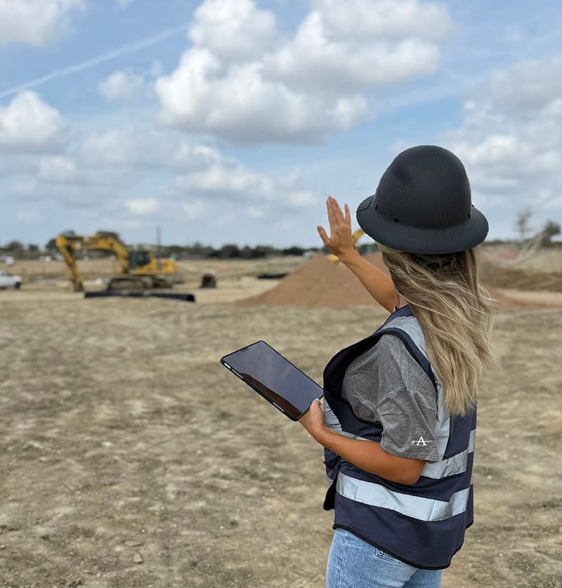 Woman in a hard-hat and reflective vest holding an iPad in one hand and the other gesturing at a construction sight.  