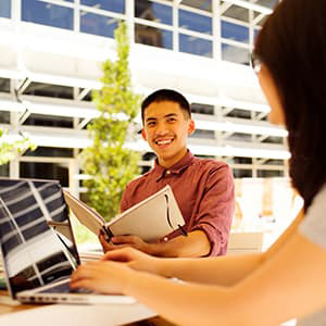 Student with notebook smiling at student with laptop