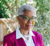 Woman with short gray hair, green-rimmed glasses, and a pink blazer with a closed-mouth smile.