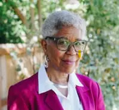 Woman with short gray hair, green-rimmed glasses, and a pink blazer with a closed-mouth smile.