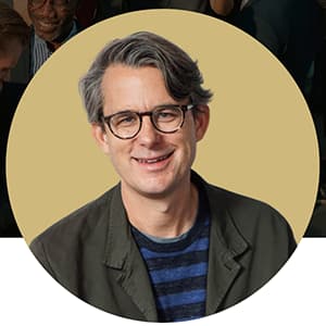 Man wearing a dark green jacket, stripped shirt, and dark-rimmed glasses is smiling.