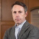 Man in a gray suit jacket and patterned white shirt with a stoic look on his face.