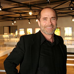 A man dressed in black and smiling with his teeth stands in a studio displaying architecture models.
