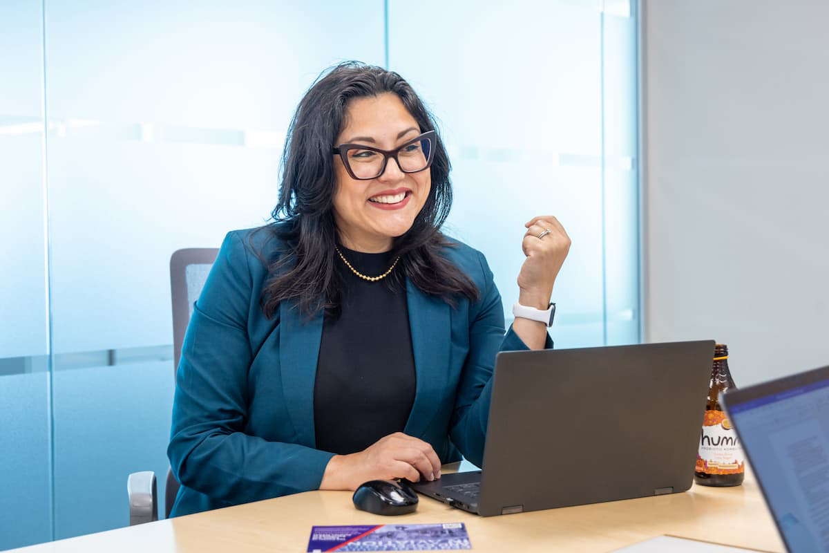 Woman with long dark hair and dark glasses smiles with her teeth as she's seated in front of a laptop computer.
