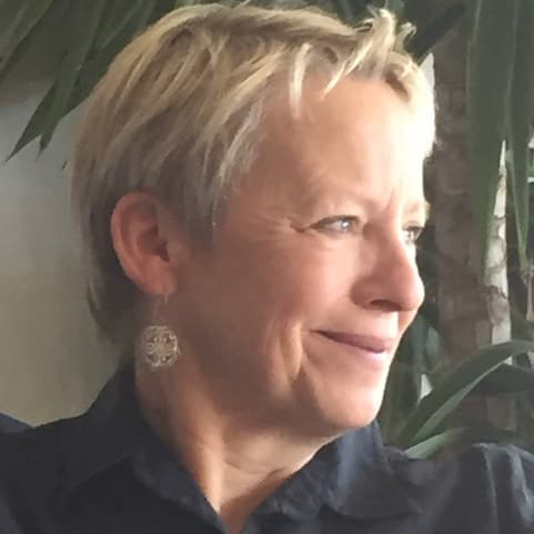 Carol Peeples with short, blonde hair looking to the side out a window, wearing a black collared shirt and earrings.
