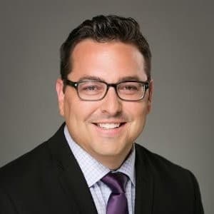Man in a suit and tie and dark rimmed glasses smiling with his teeth.