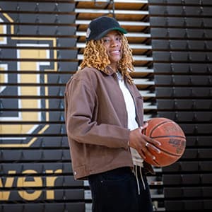 Woman in a black baseball cap and brown jacket holds a basketball and smiles with her teeth.