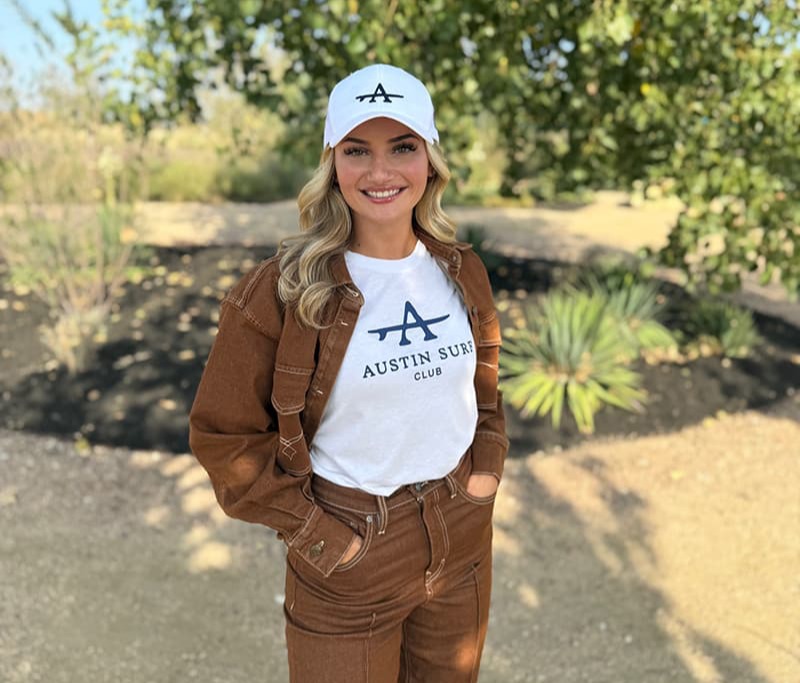 Woman with long blonde hair smiling with her teeth wearing a white Austin Surf Club hat and t-shirt.