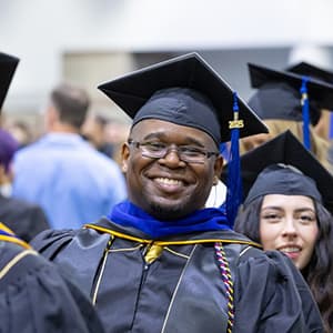 Black man wearing commencement regalia and glasses smiles with his teeth.