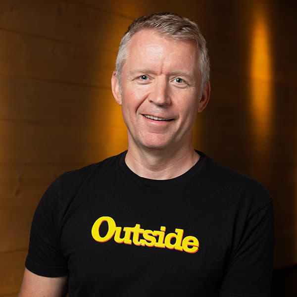 Robin Thurston wearing a black t-shirt with "Outside" in yellow writing across the front. He has short gray hair and giving a side smile with his teeth.