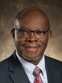 Headshot of Dr. Sam Dosumu in a dark suit and colorful tie with glasses and a closed-mouth smile.