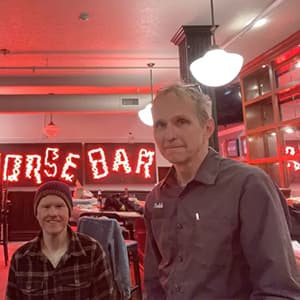 Two men smiling in a bar with a red neon sign behind them on the wall.