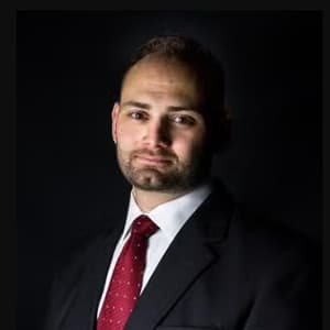 Man in a dark suit and red tie against a black background and a closed-mouth smile.