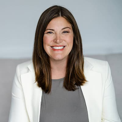 Kristin Wegner Guilfoyle with mid-shoulder lengthed, dark brown hair, wearing a white suite jacket and gray shirt, smiling with her teeth.