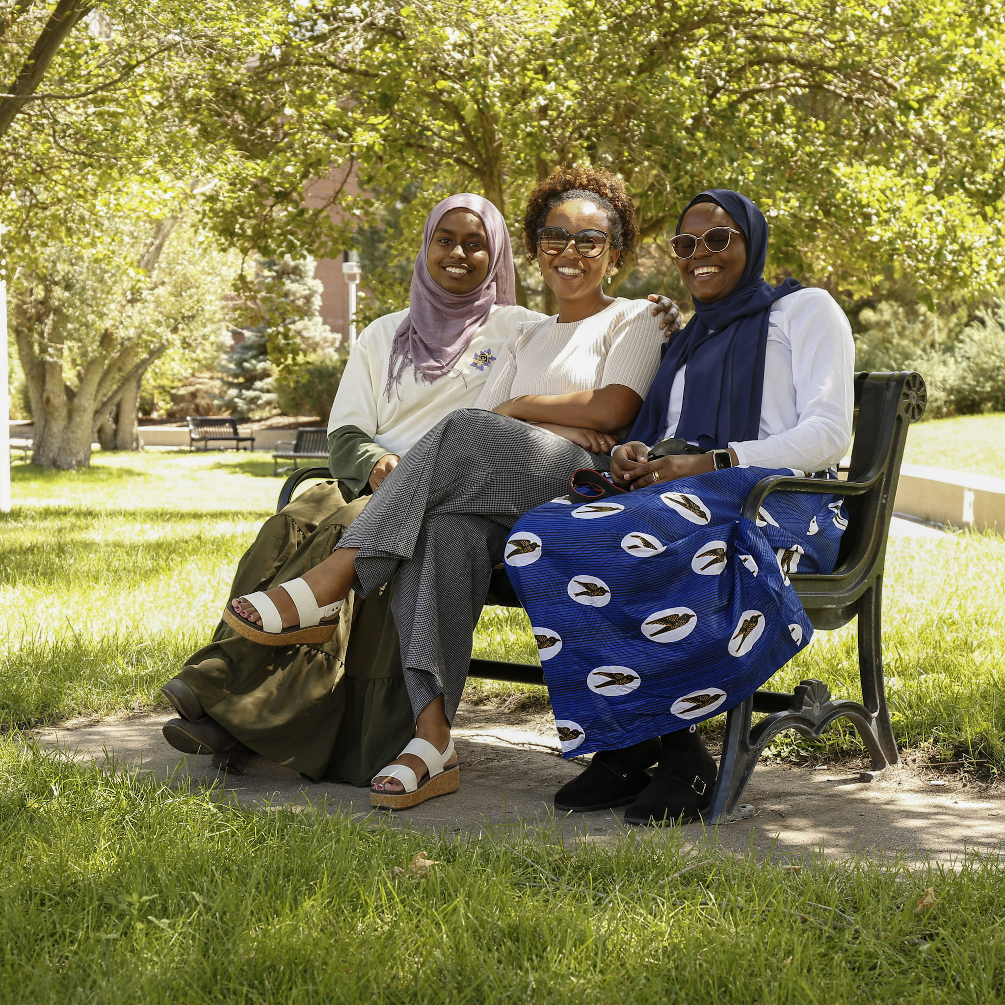 Three women sit together on a shaded bench surrounded by green grass and trees.