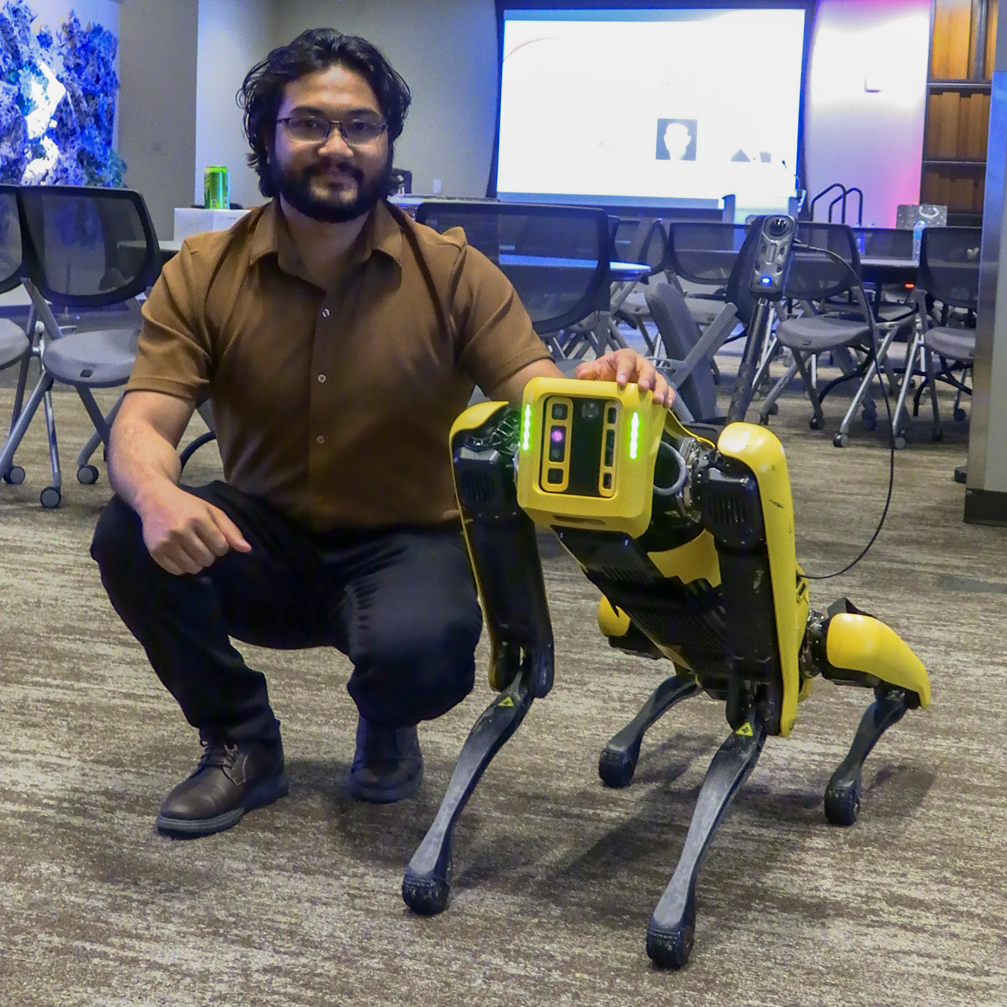 A man kneels beside Spot, a yellow quadruped robot dog, at the AI4Infrastructures Symposium.