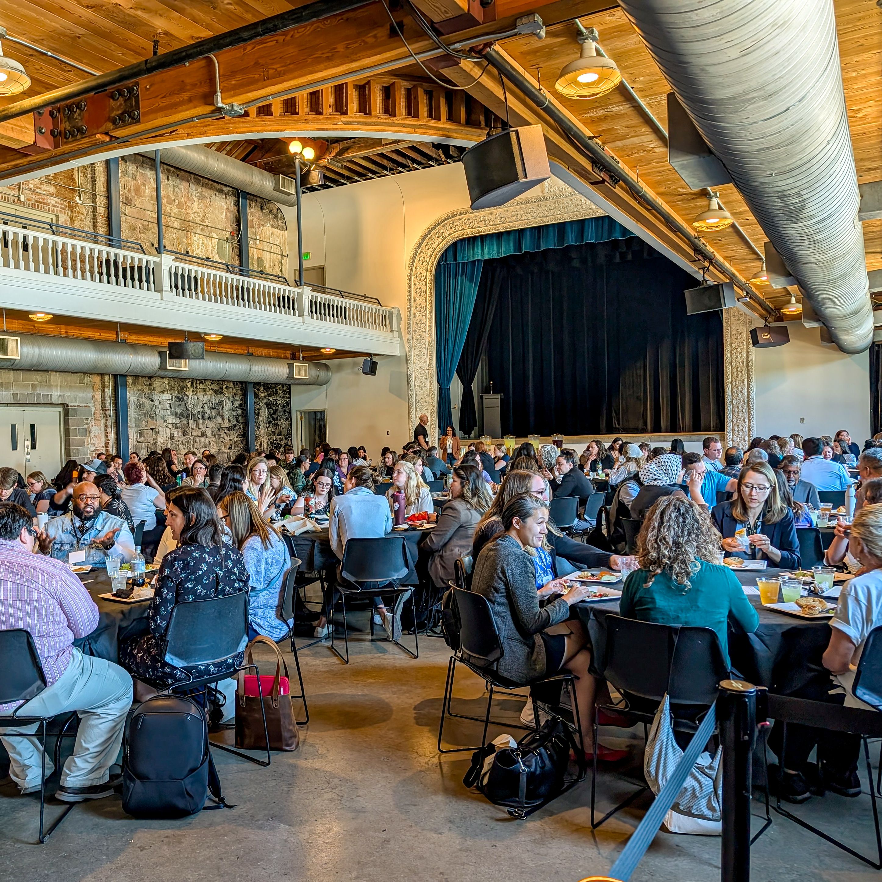 Faculty, staff, and students sit together at tables throughout the venue during the CU Inclusive Excellence Summit, sharing a meal beneath the stage and balcony