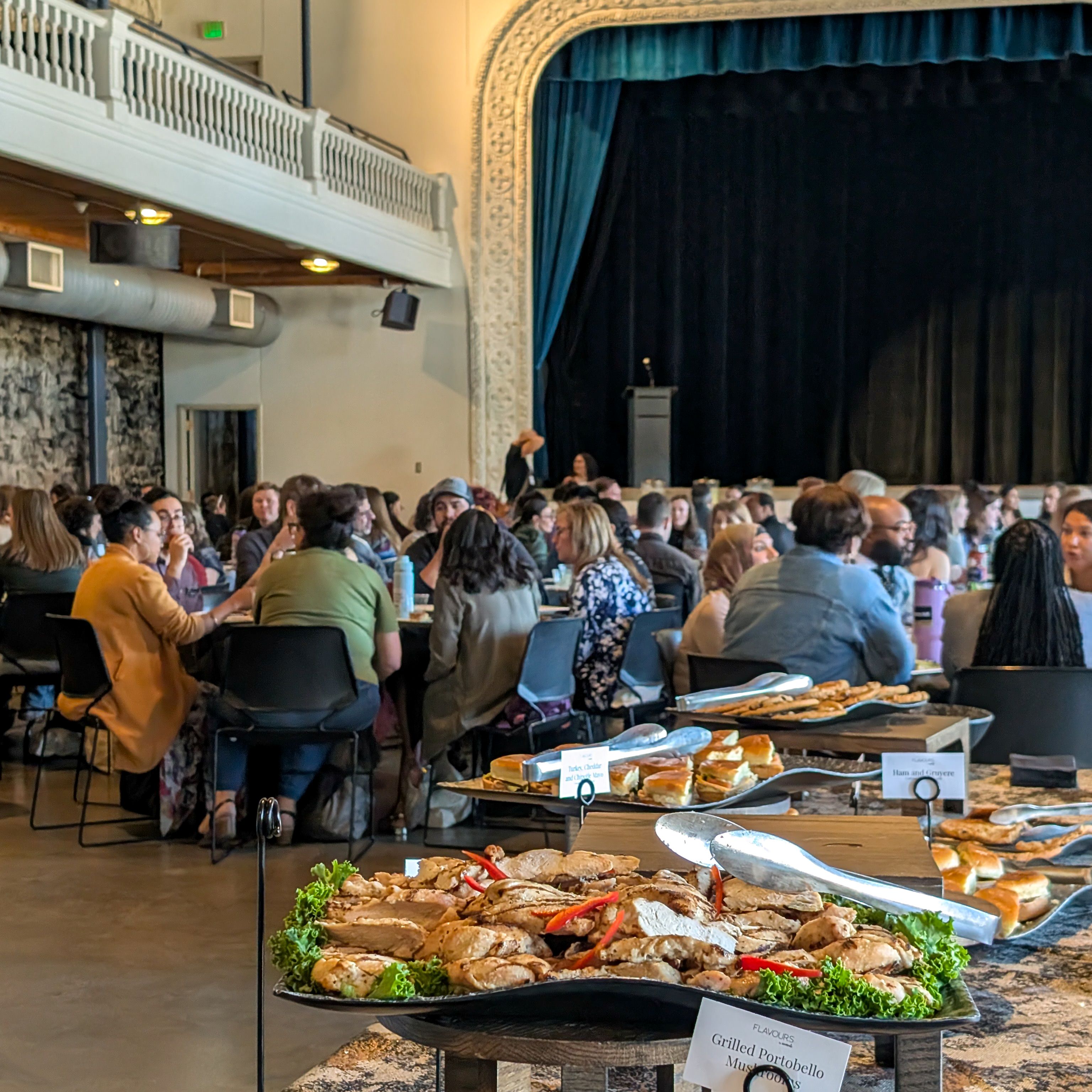 Attendees gather around tables for the CU Inclusive Excellence Summit, with a buffet of food set up in front of the stage