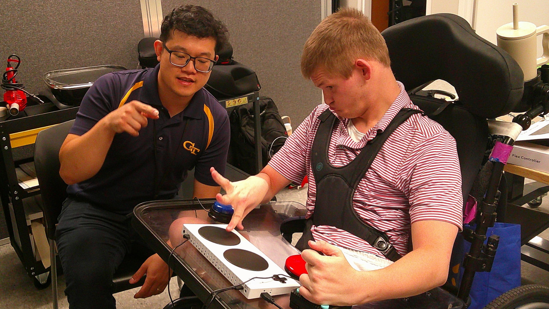 A man in glasses talks with a young man in a power wheelchair, who presses one of two large round buttons on an adaptive game controller resting on his wheelchair tray