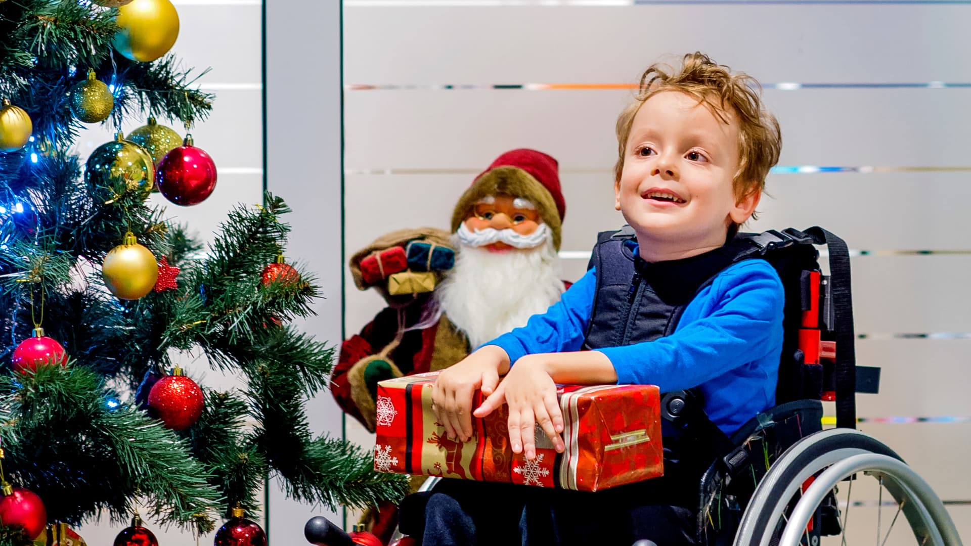 A smiling young boy in a wheelchair holds a wrapped red present on his lap, next to a decorated Christmas tree and a Santa Claus figure