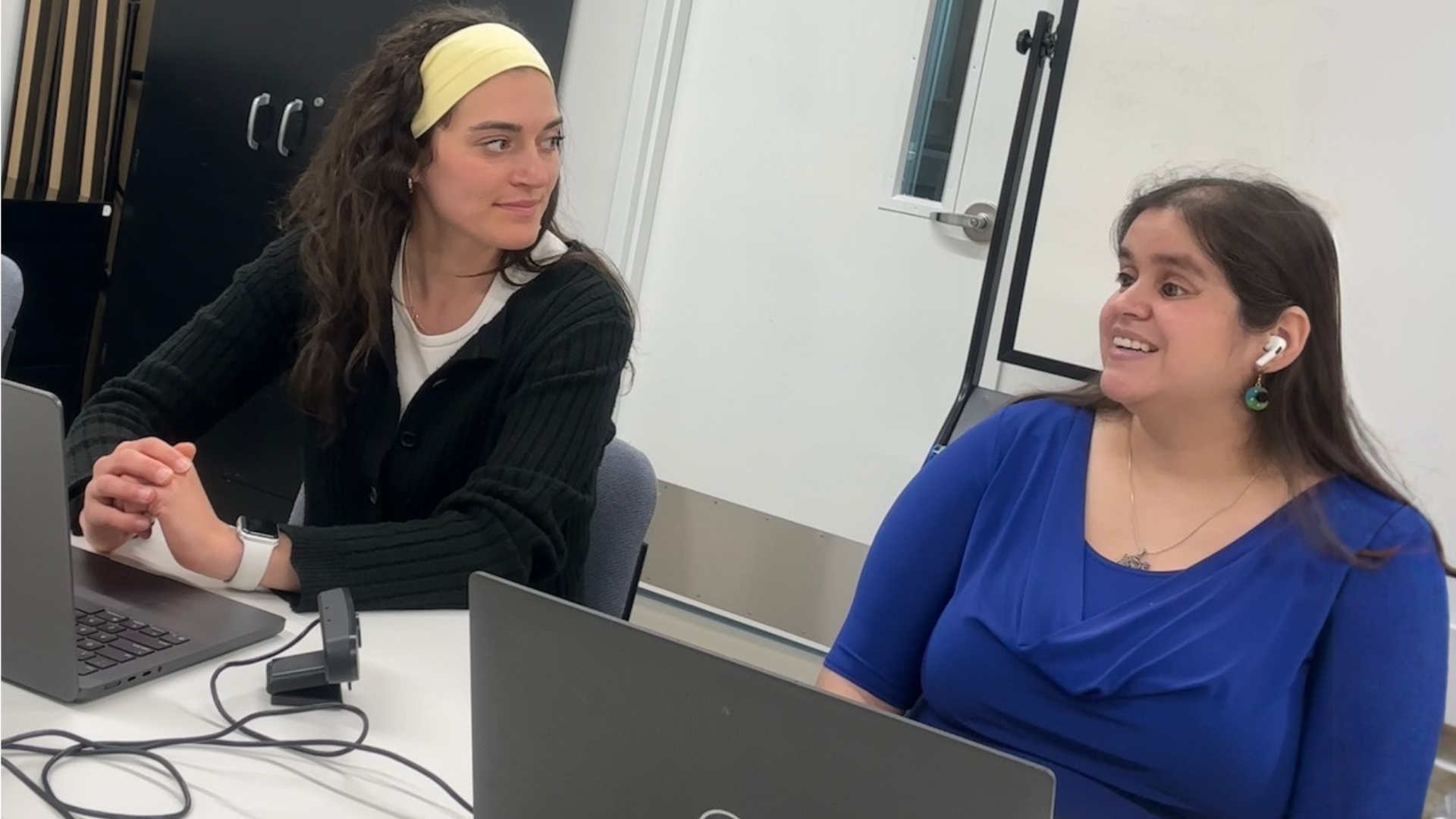 Two women sit at a table with laptops. One, wearing earbuds, smiles as she talks while the other listens.