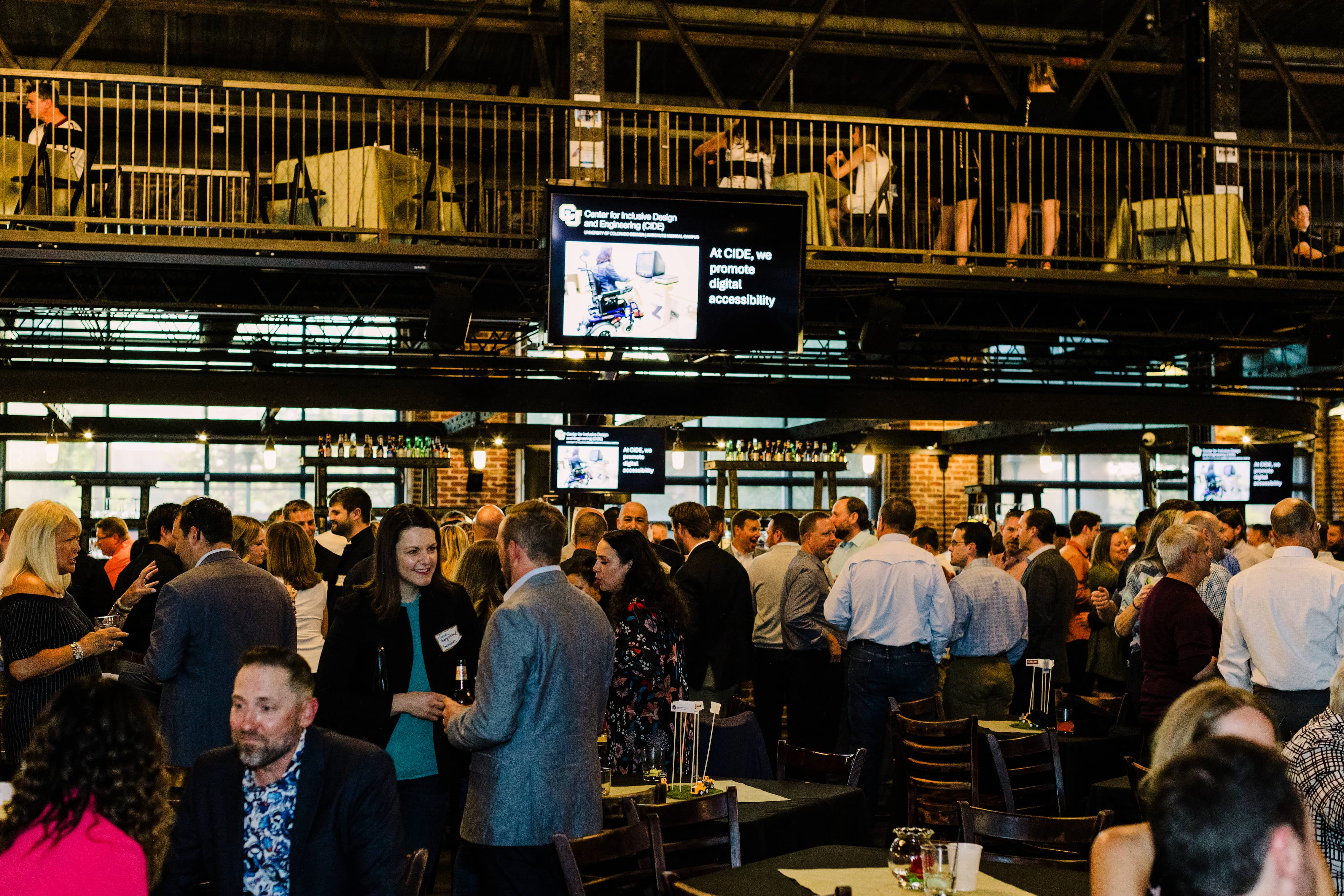 A large group in business casual in a softly lit two story venue. An overhead TV shows the CIDE logo and the text at CIDE, we promote digital accessibility.