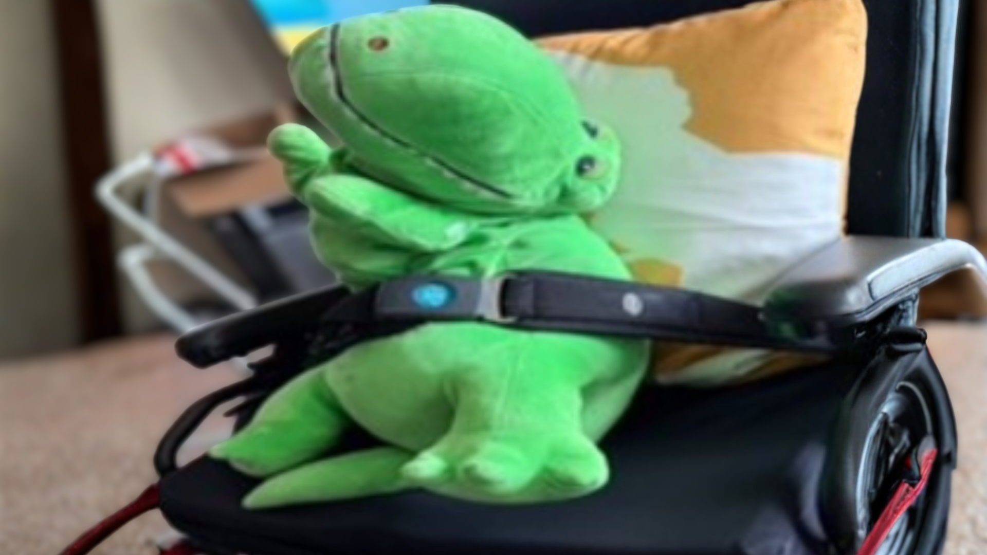 A green stuffed dinosaur sits buckled into a wheelchair seat with a lap belt, propped against a yellow and white pillow.