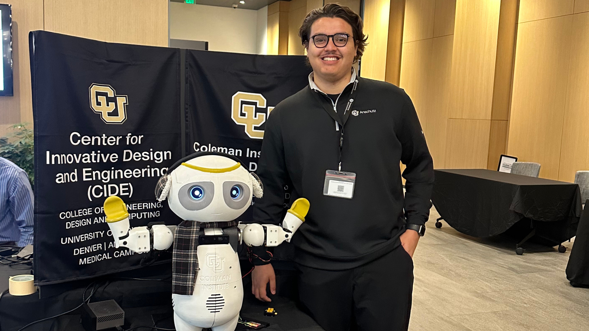 A smiling young man in glasses and a black quarter-zip stands beside a small white robot with large blue eyes and a plaid scarf, in front of CIDE and Coleman Institute banners.