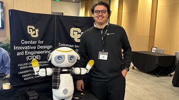 A smiling young man in glasses and a black quarter-zip stands beside a small white robot with large blue eyes and a plaid scarf, in front of CIDE and Coleman Institute banners.