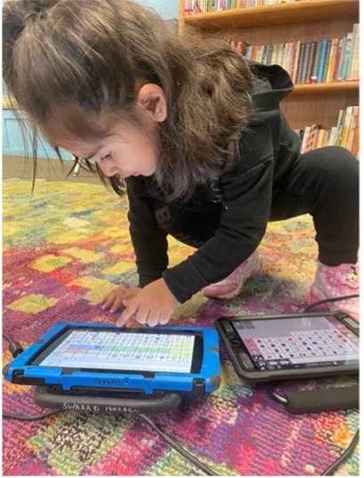 young girl crouching over two AAC tablet-based devices, pointing towards a word on the screen
