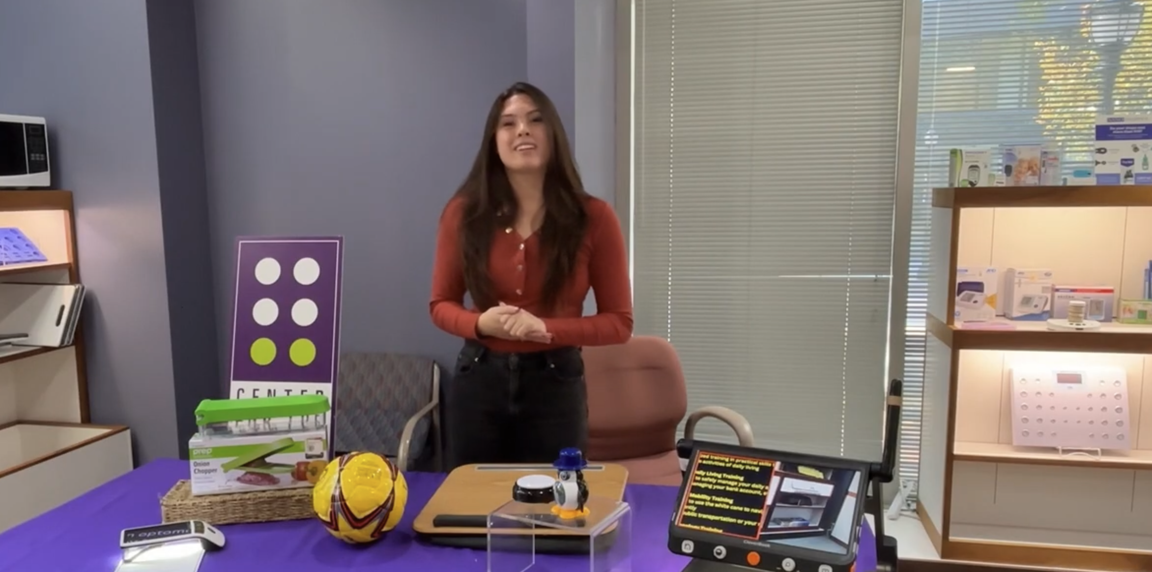 A smiling woman stands behind a table covered in gifts for people with visual impairment.